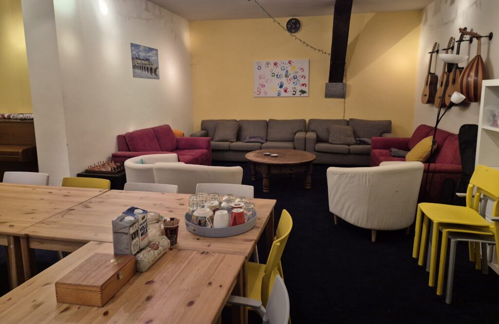 De ruimte van het café. Op de voorgrond een lange houten tafel met stoelen, daarachter een zithoek met banken en fauteuils.